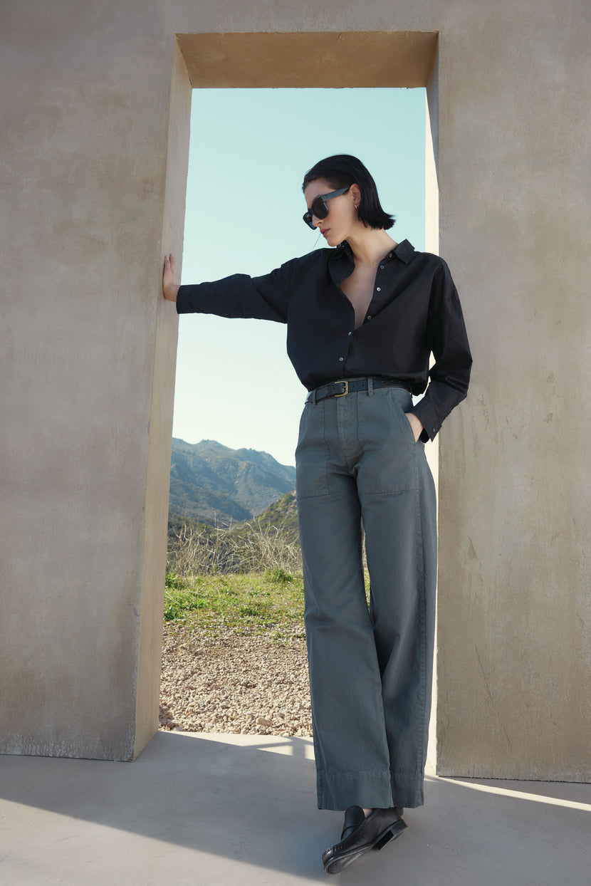 A person wearing a black shirt and Velvet by Jenny Graham's VENTURA PANT leans against a doorway, with mountains visible in the background. The individual has short dark hair and is sporting sunglasses.