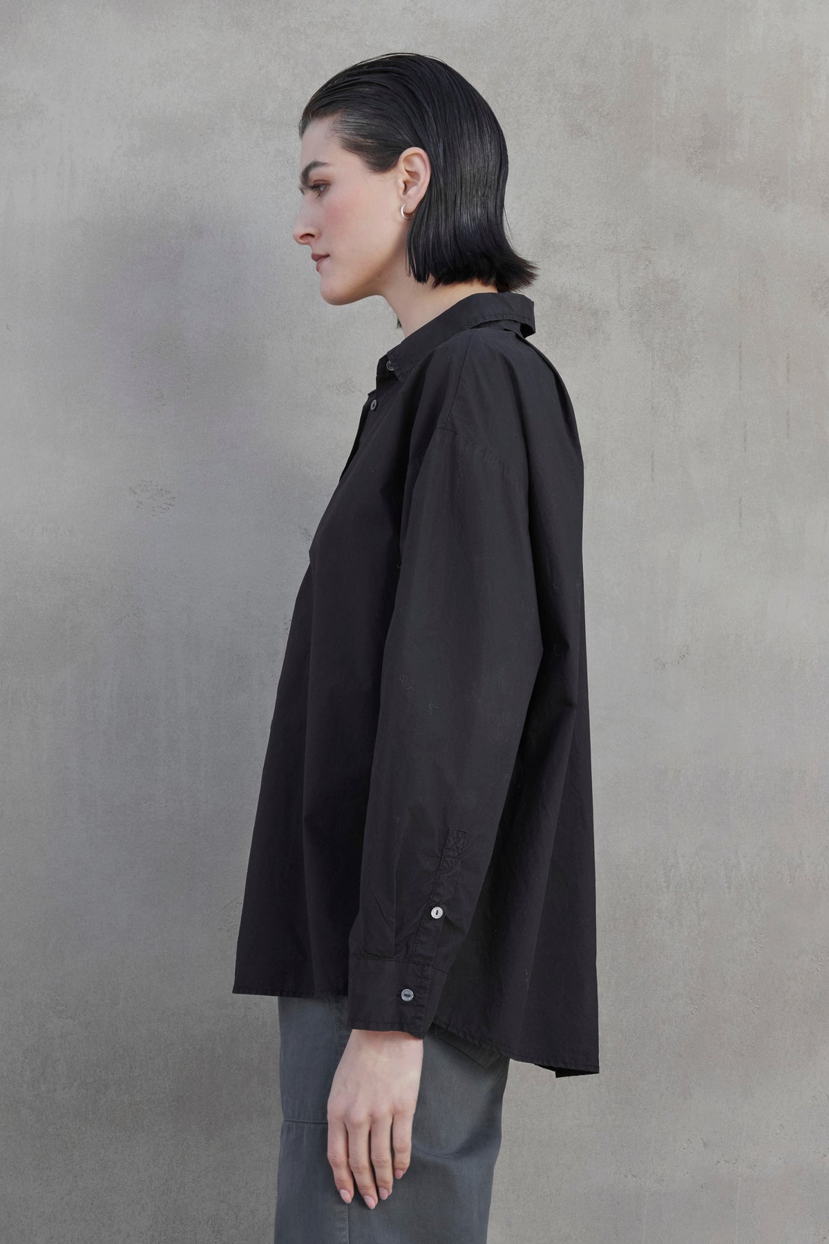   A person with short, slicked-back hair wearing a black DAKOTA SHIRT by Velvet by Jenny Graham stands in profile against a grey, textured background. 