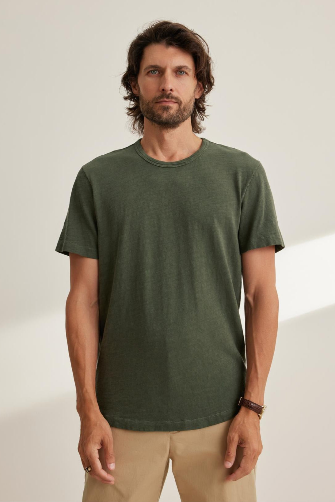   A man with medium-length hair and a beard is wearing the SPADER TEE, a relaxed fit green Peruvian cotton slub t-shirt by Velvet by Graham & Spencer, paired with beige pants. He stands against a plain background. 