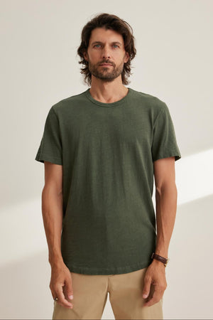 A man with medium-length hair and a beard is wearing the SPADER TEE, a relaxed fit green Peruvian cotton slub t-shirt by Velvet by Graham & Spencer, paired with beige pants. He stands against a plain background.