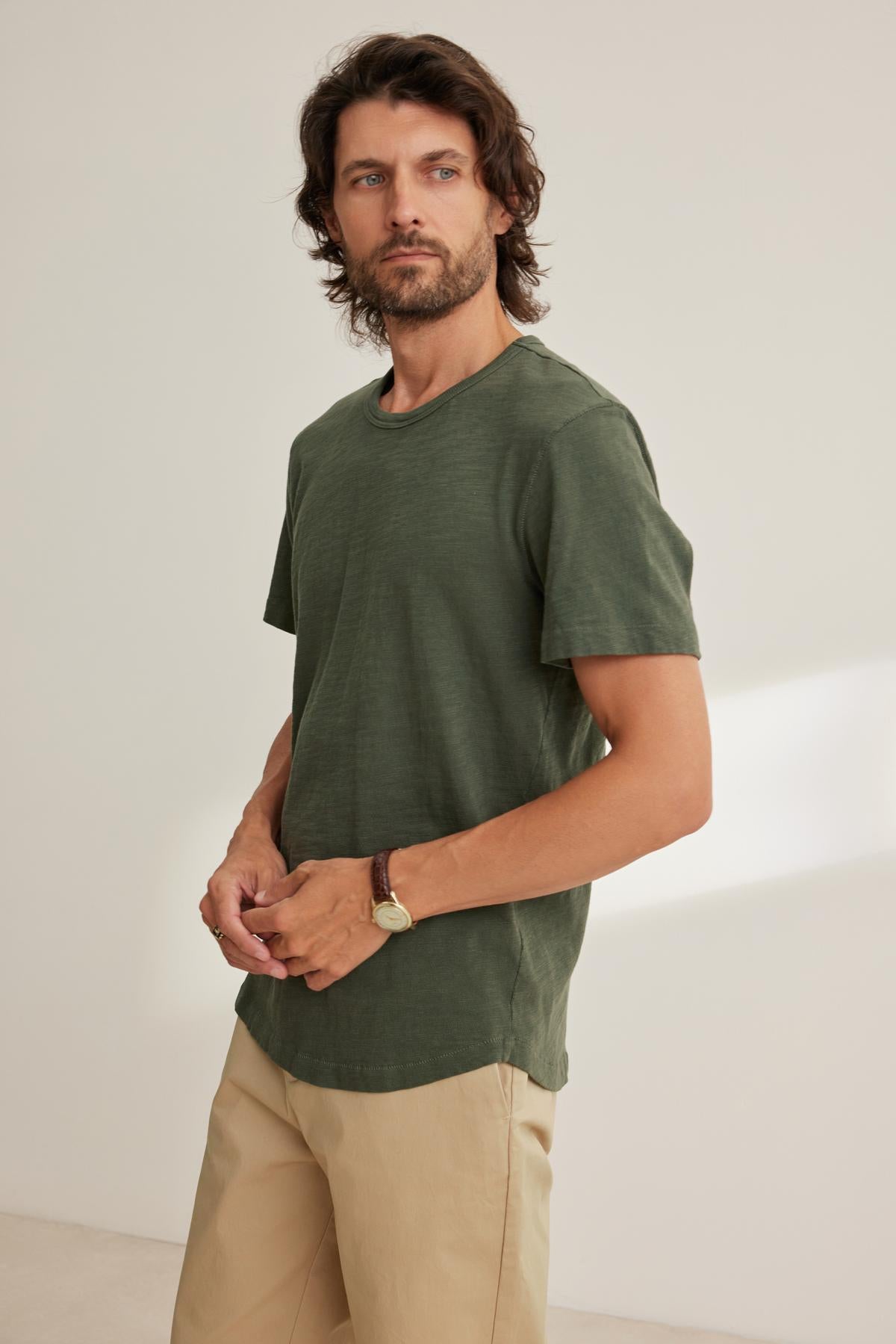 A man with medium-length hair wears a SPADER TEE by Velvet by Graham & Spencer, featuring a classic crew neckline and relaxed fit in green, paired with beige pants. Crafted from mid-weight Peruvian cotton, the outfit enhances his easygoing style against the plain background.-38643352699073