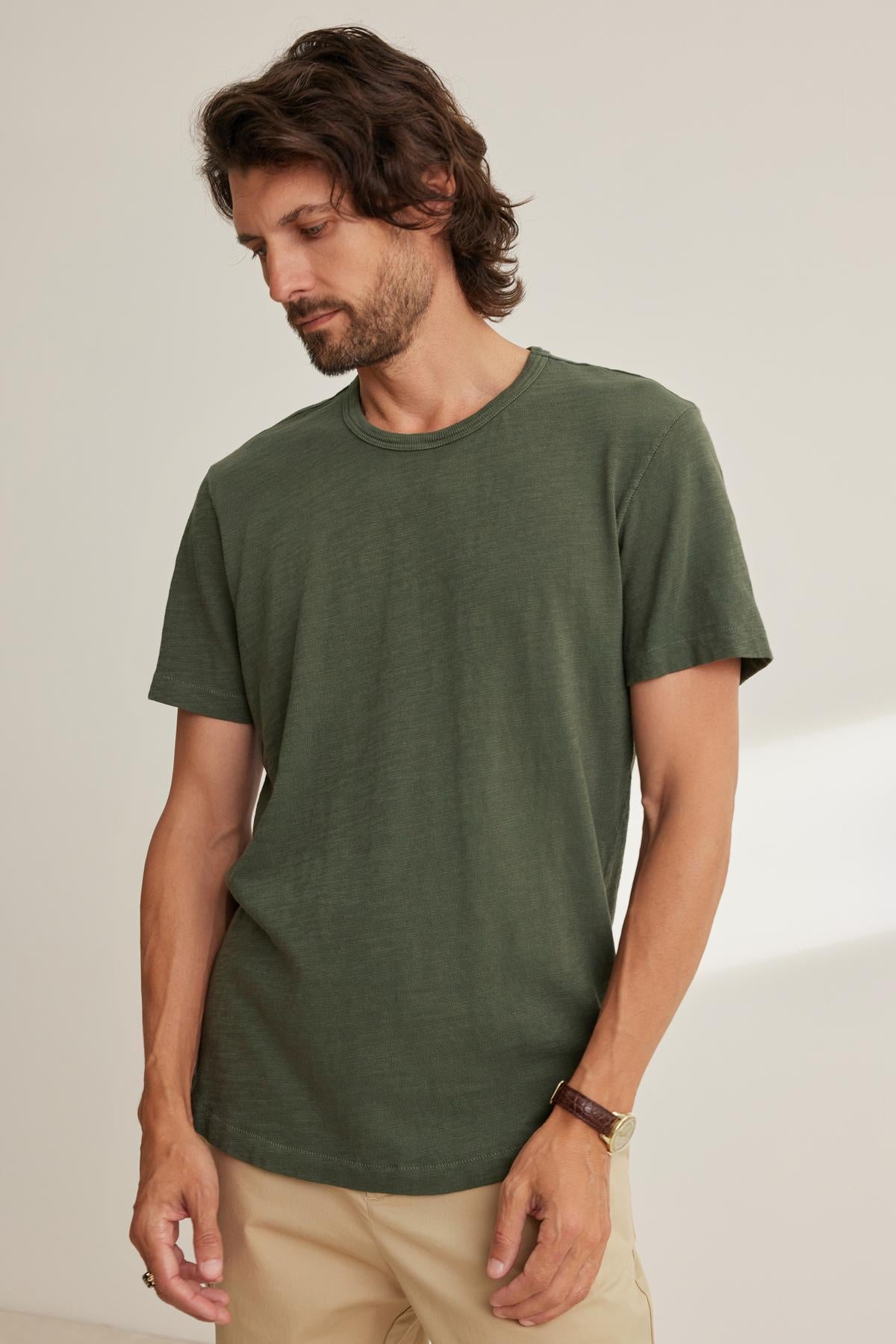   A person with medium-length hair is standing indoors, looking down. They are wearing the SPADER TEE from Velvet by Graham & Spencer, a green t-shirt featuring a classic crew neckline, and beige pants. 