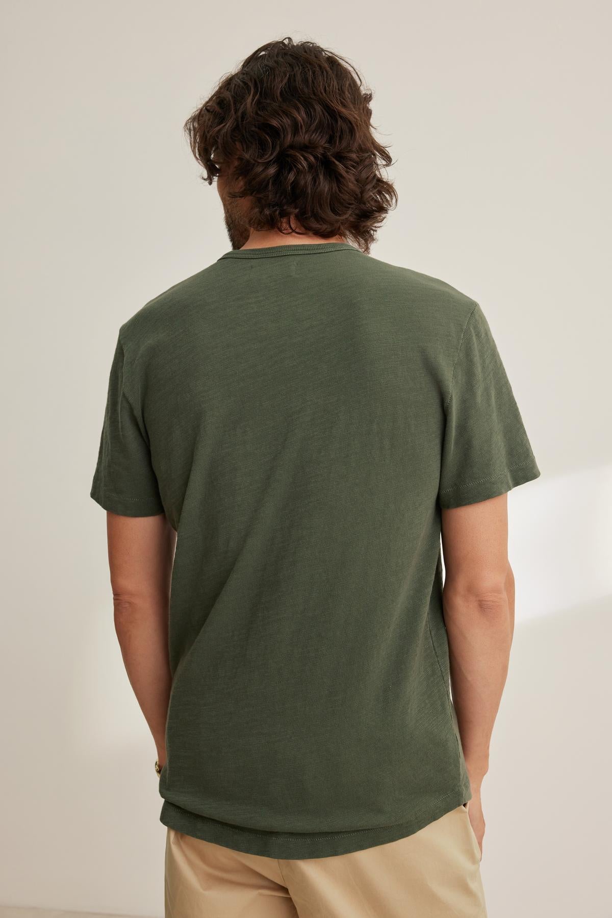 A person with medium-length hair faces away, wearing a Velvet by Graham & Spencer SPADER TEE in dark green with a classic crew neckline and beige pants. The relaxed fit of the mid-weight Peruvian cotton tee complements the softly lit room's ambiance.-38643352797377