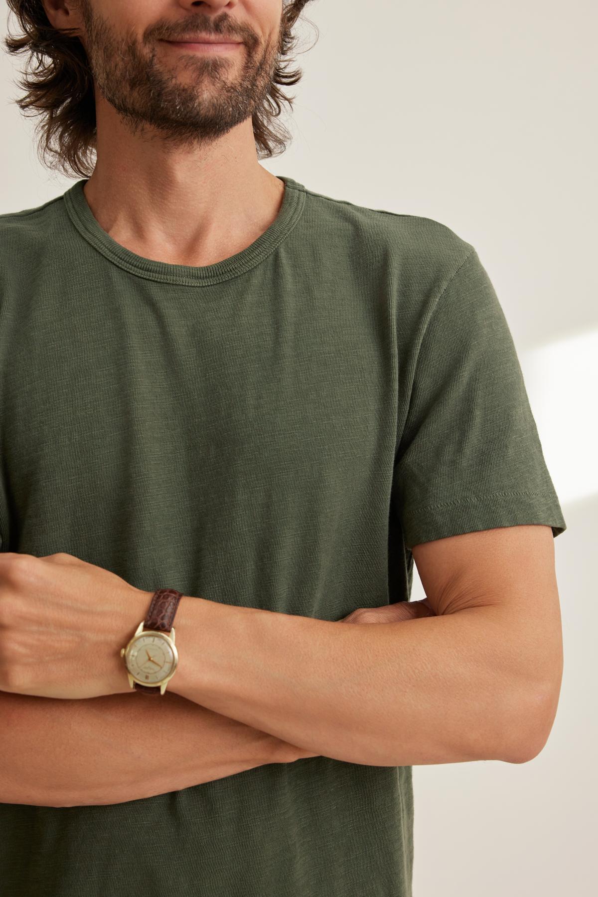   A person in a relaxed fit green SPADER TEE from Velvet by Graham & Spencer crosses their arms, displaying a gold wristwatch with a brown leather strap. 