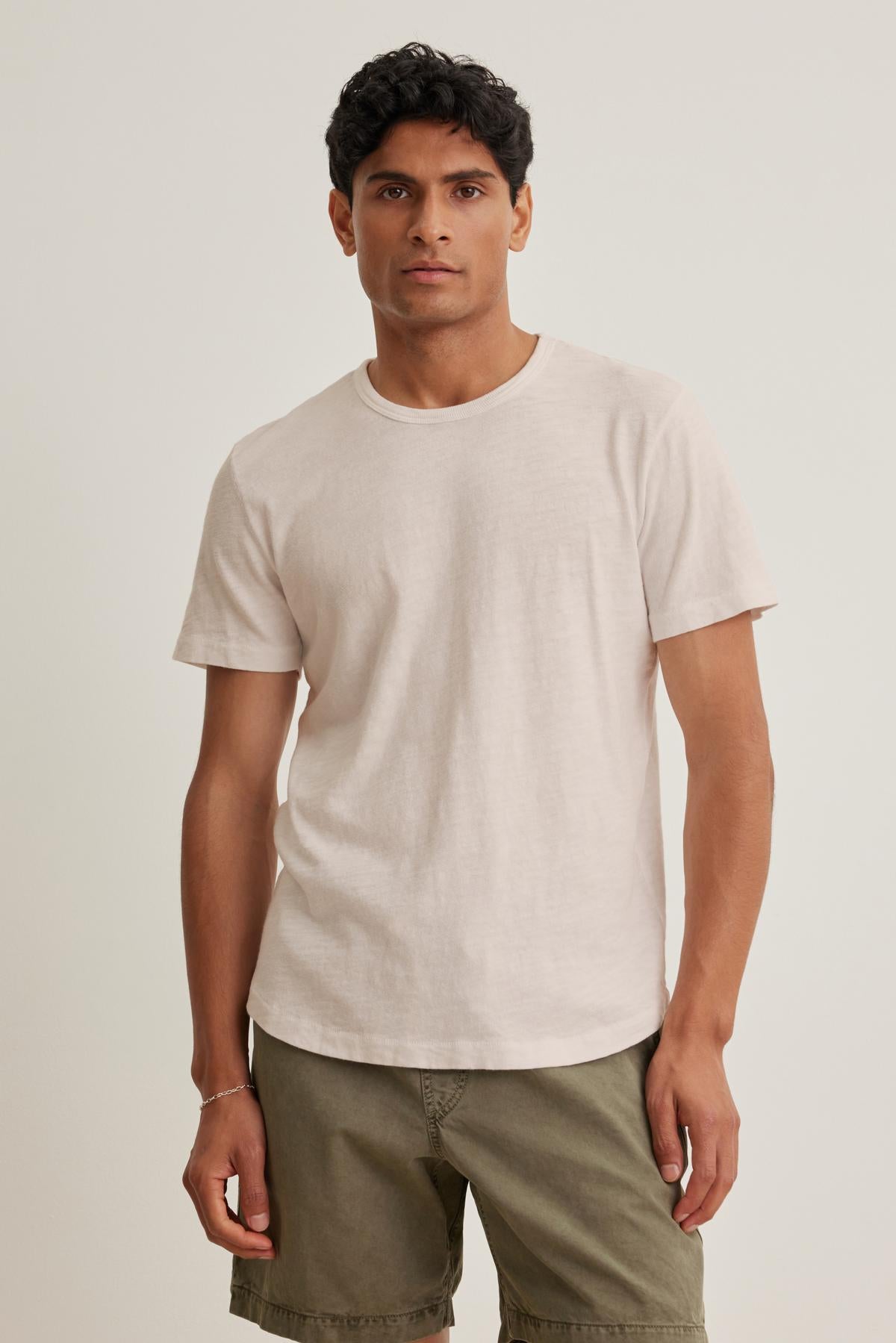 A person stands against a neutral background wearing the laid-back SPADER TEE by Velvet by Graham & Spencer, a light beige t-shirt crafted from mid-weight Peruvian cotton, paired with khaki shorts.-38658664300737