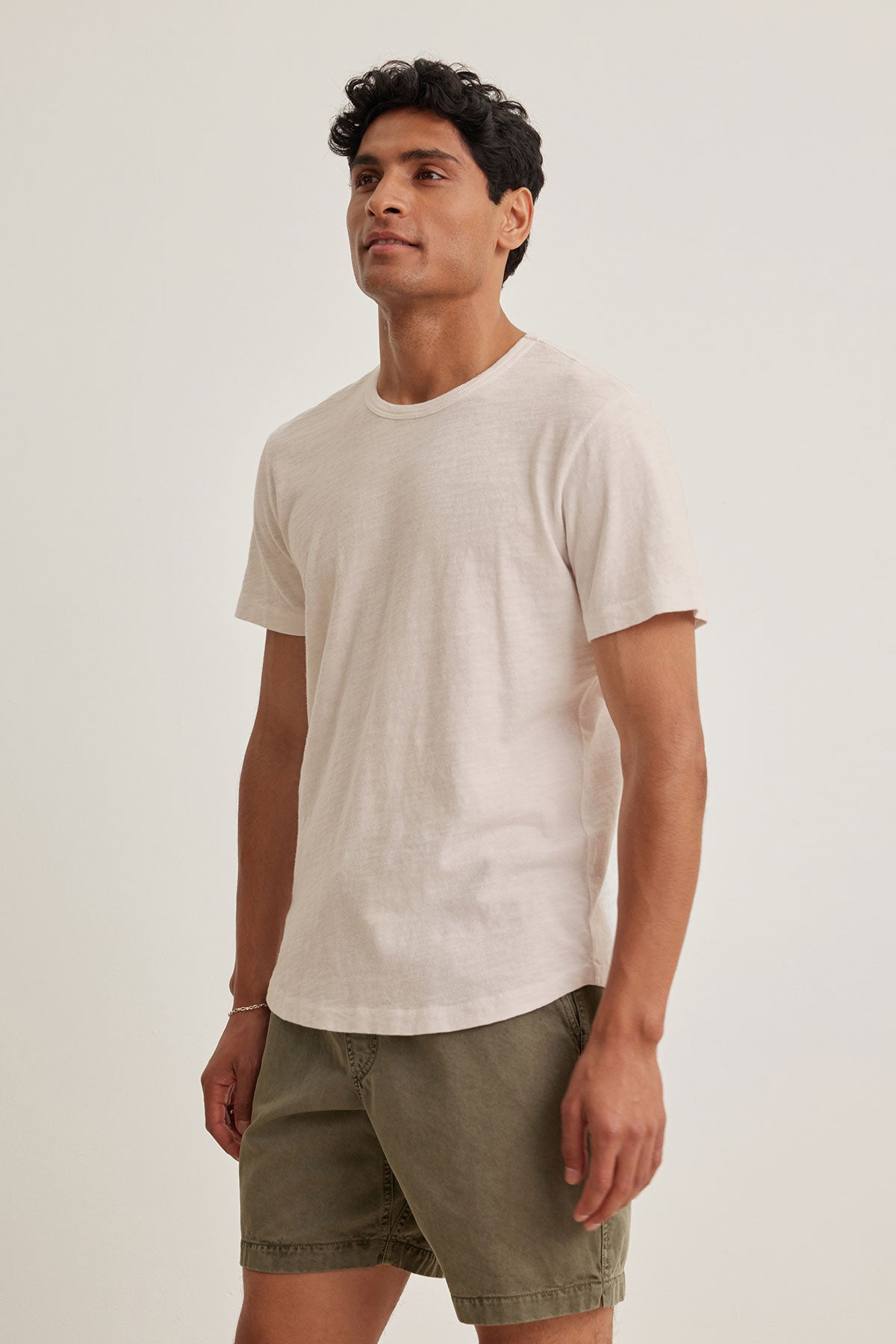   A person wearing a SPADER TEE by Velvet by Graham & Spencer made of mid-weight Peruvian cotton, in light beige with a classic crew neck, along with olive green shorts, stands against a plain white background. 