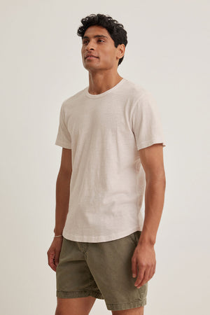 A person wearing a SPADER TEE by Velvet by Graham & Spencer made of mid-weight Peruvian cotton, in light beige with a classic crew neck, along with olive green shorts, stands against a plain white background.