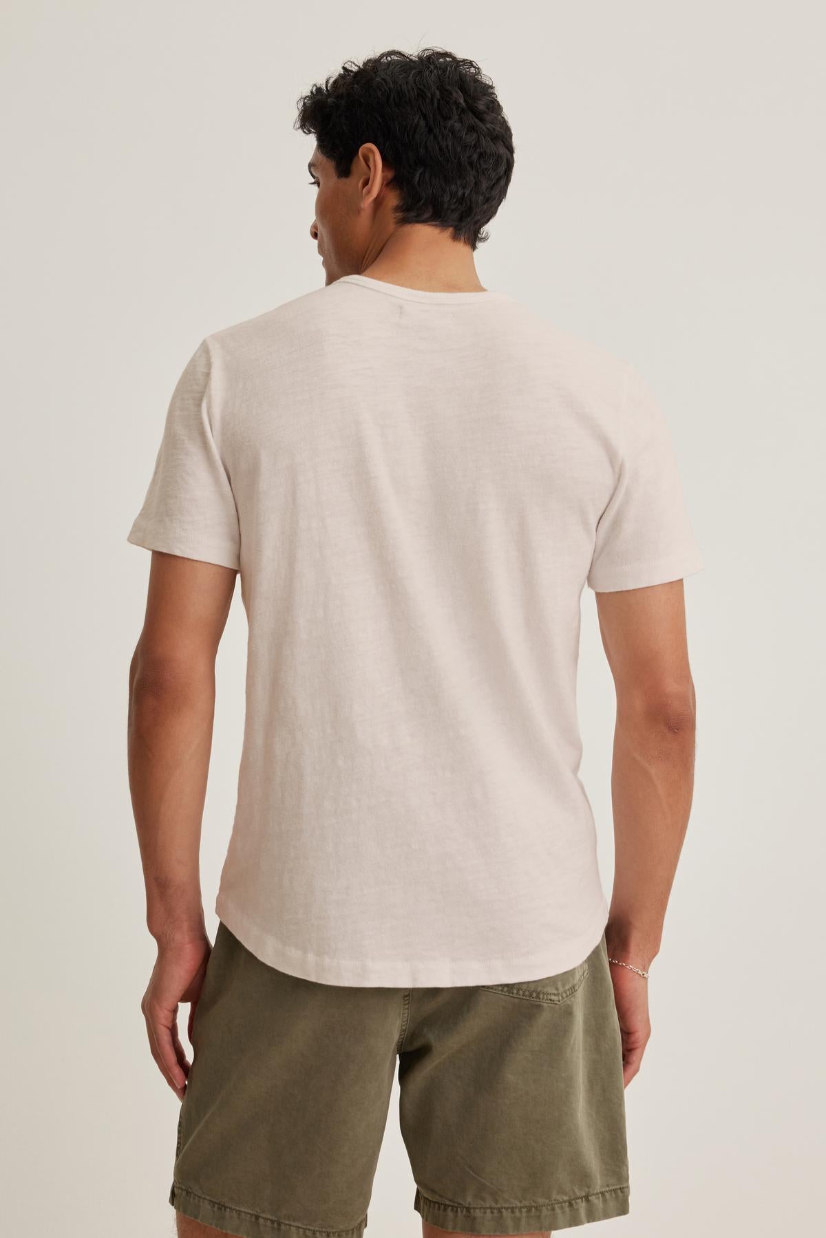 A laid-back man in the SPADER TEE by Velvet by Graham & Spencer, a relaxed-fit mid-weight Peruvian cotton white t-shirt, and olive shorts is seen from behind against a plain backdrop.-38658664333505