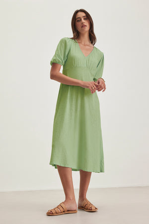 Barefoot in her light green PARKER DRESS by Velvet by Graham & Spencer, a woman stands against a plain white background and light-colored floor. The figure-skimming cotton slub dress enhances her effortless style.