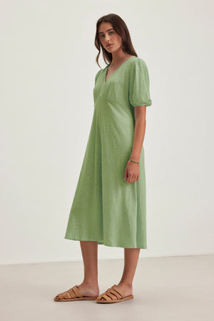 A woman in the Velvet by Graham & Spencer PARKER DRESS, a green figure-skimming midi dress, and brown sandals stands against a plain background.