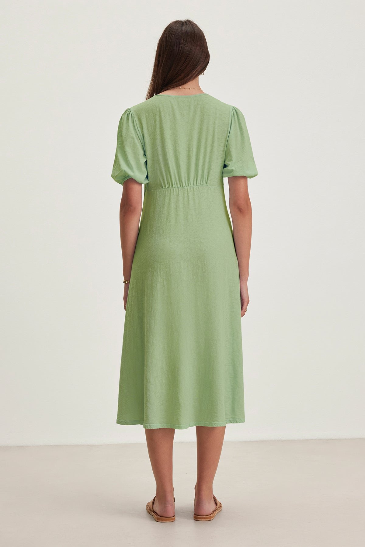   Rear view of a person with long hair in the PARKER DRESS by Velvet by Graham & Spencer, a figure-skimming cotton slub dress in light green, paired with flat sandals on a light-colored surface. 