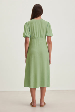 Rear view of a person with long hair in the PARKER DRESS by Velvet by Graham & Spencer, a figure-skimming cotton slub dress in light green, paired with flat sandals on a light-colored surface.