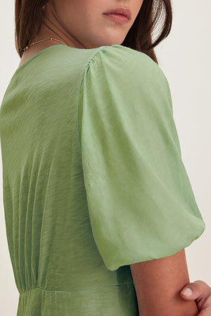 A woman is pictured from the back and side, wearing the PARKER DRESS by Velvet by Graham & Spencer, a light green blouse with puffed sleeves and a figure-skimming fit.