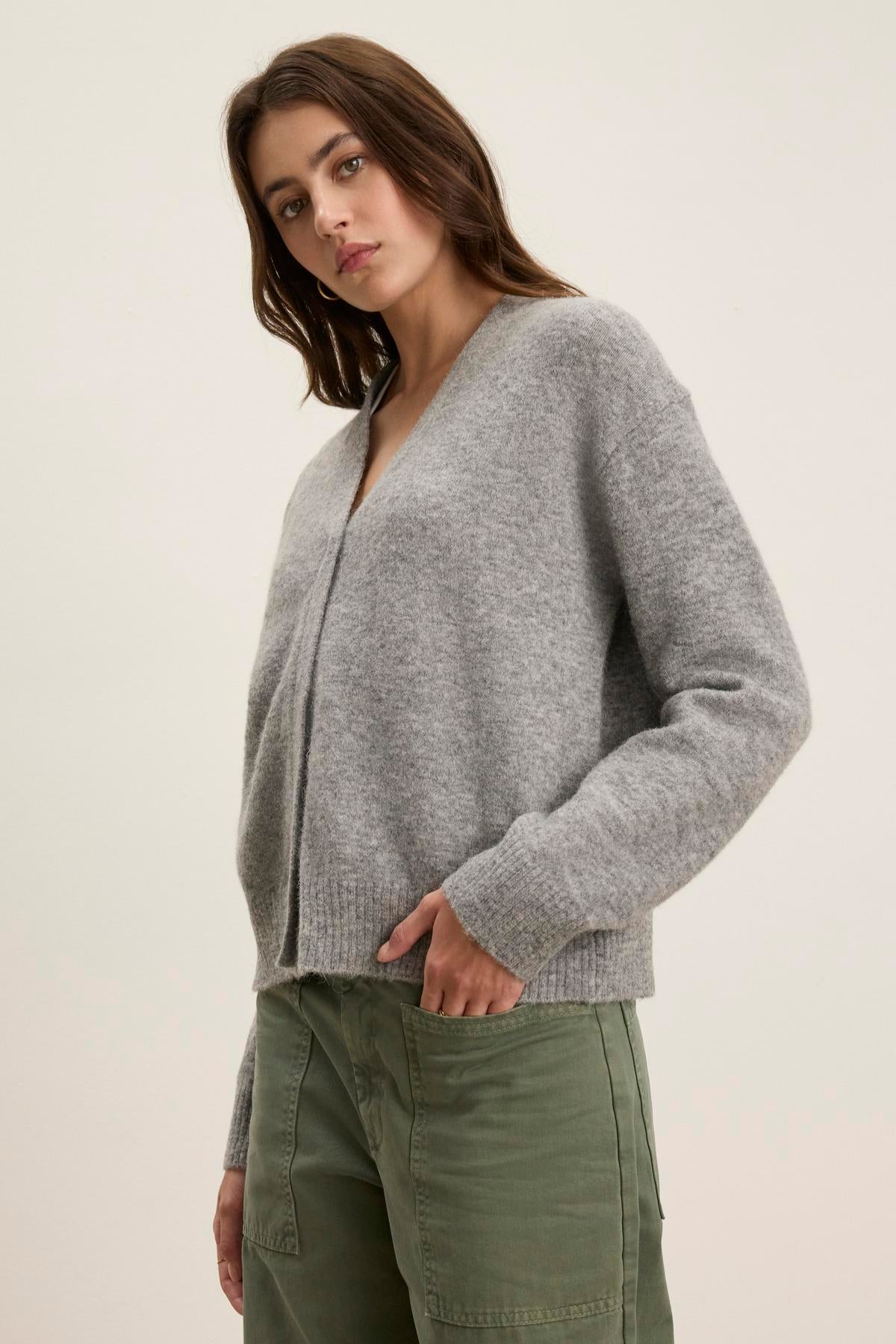 A person in a Velvet by Graham & Spencer MARTI ALPACA CARDIGAN with ribbed trims and green pants stands against a plain backdrop, gazing at the camera with a neutral expression.-38513426628801