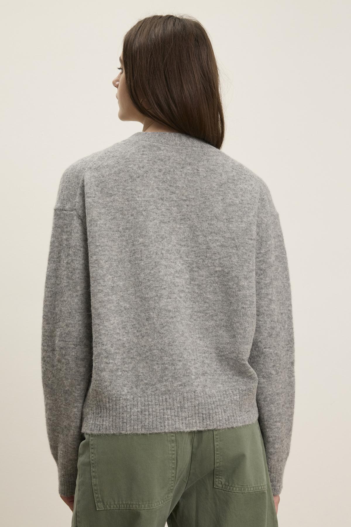   Someone with long dark hair, wearing the MARTI ALPACA CARDIGAN by Velvet by Graham & Spencer and green pants, faces away from the camera against a pale background, highlighting the elegant ribbed trims. 