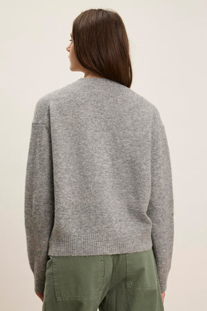 Someone with long dark hair, wearing the MARTI ALPACA CARDIGAN by Velvet by Graham & Spencer and green pants, faces away from the camera against a pale background, highlighting the elegant ribbed trims.