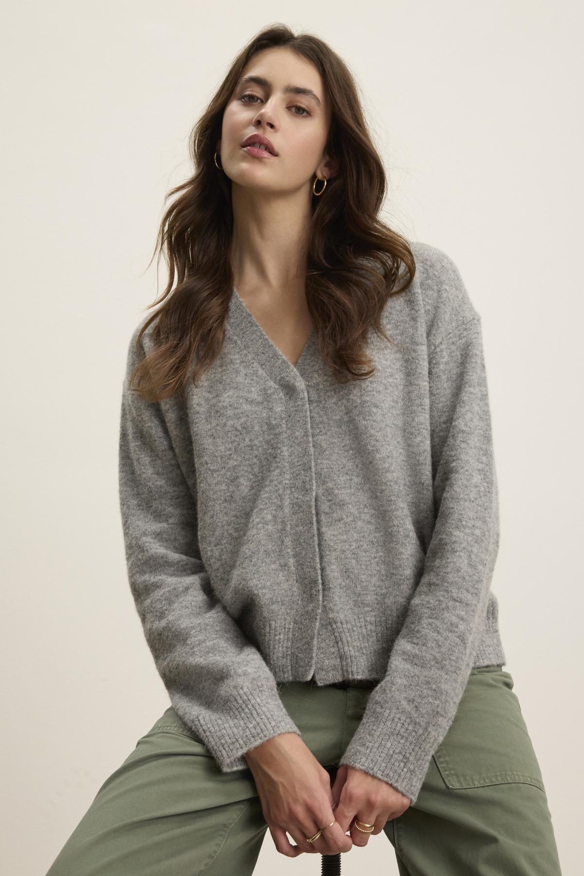   A person with long brown hair is wearing the MARTI ALPACA CARDIGAN by Velvet by Graham & Spencer, a cozy gray sweater with ribbed trims, and green pants, posing against a plain background. 