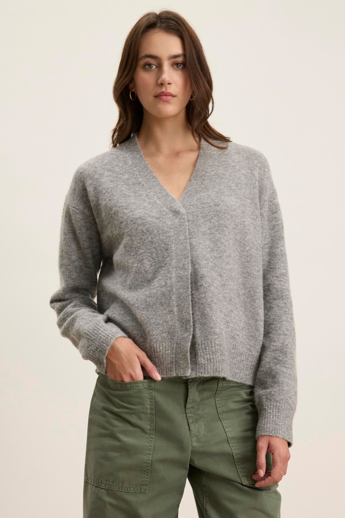 A woman in a gray Velvet by Graham & Spencer MARTI ALPACA CARDIGAN and green pants stands against a plain background.-38513426727105