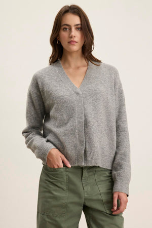 A woman in a gray Velvet by Graham & Spencer MARTI ALPACA CARDIGAN and green pants stands against a plain background.