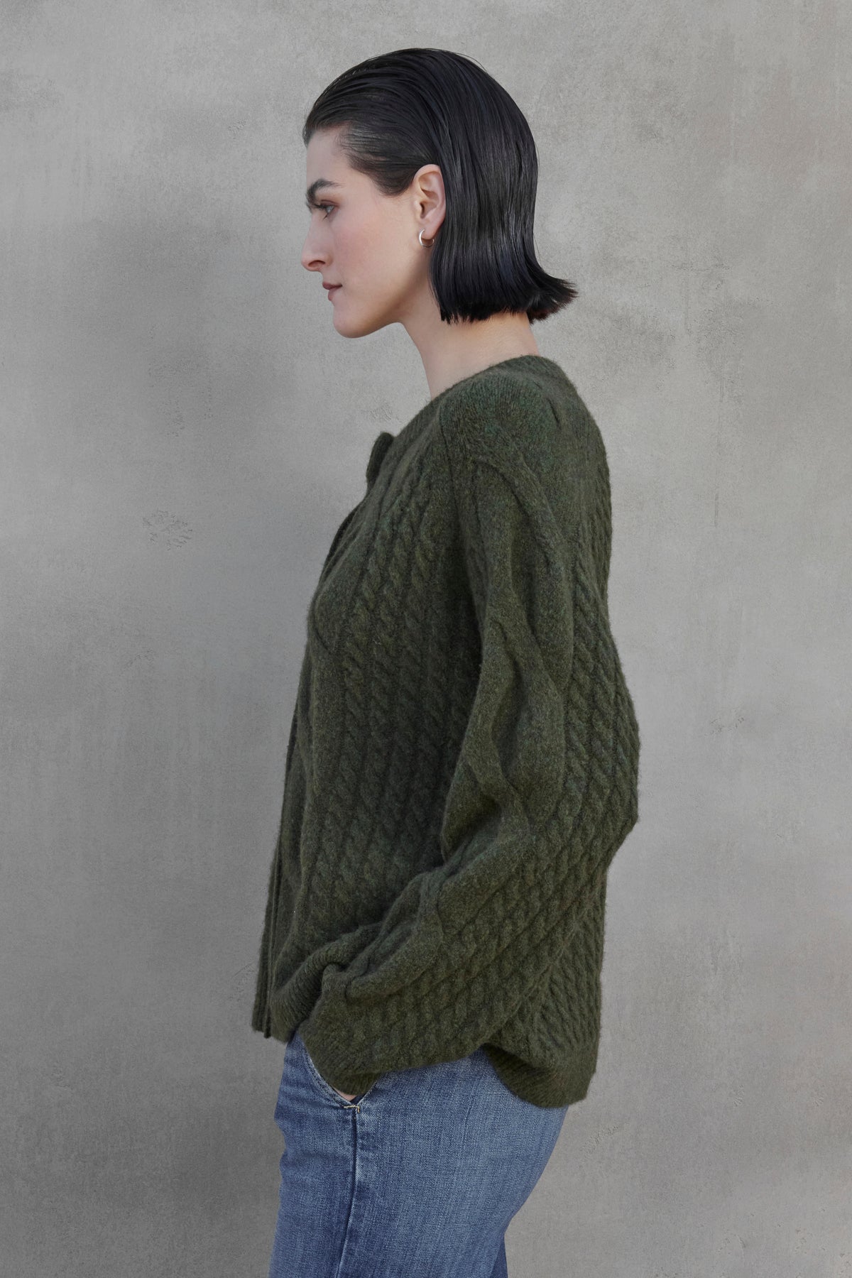   A person with short dark hair is shown in profile wearing the BEACHWOOD CARDIGAN by Velvet by Jenny Graham, paired with blue jeans, standing against a gray background. 