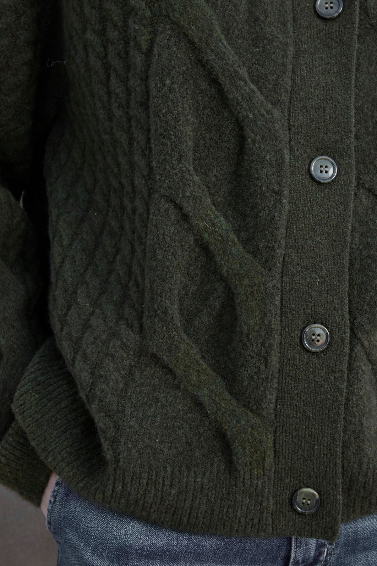   Close-up of the BEACHWOOD CARDIGAN, a dark green cable knit piece by Velvet by Jenny Graham, featuring black buttons and paired with blue jeans. 