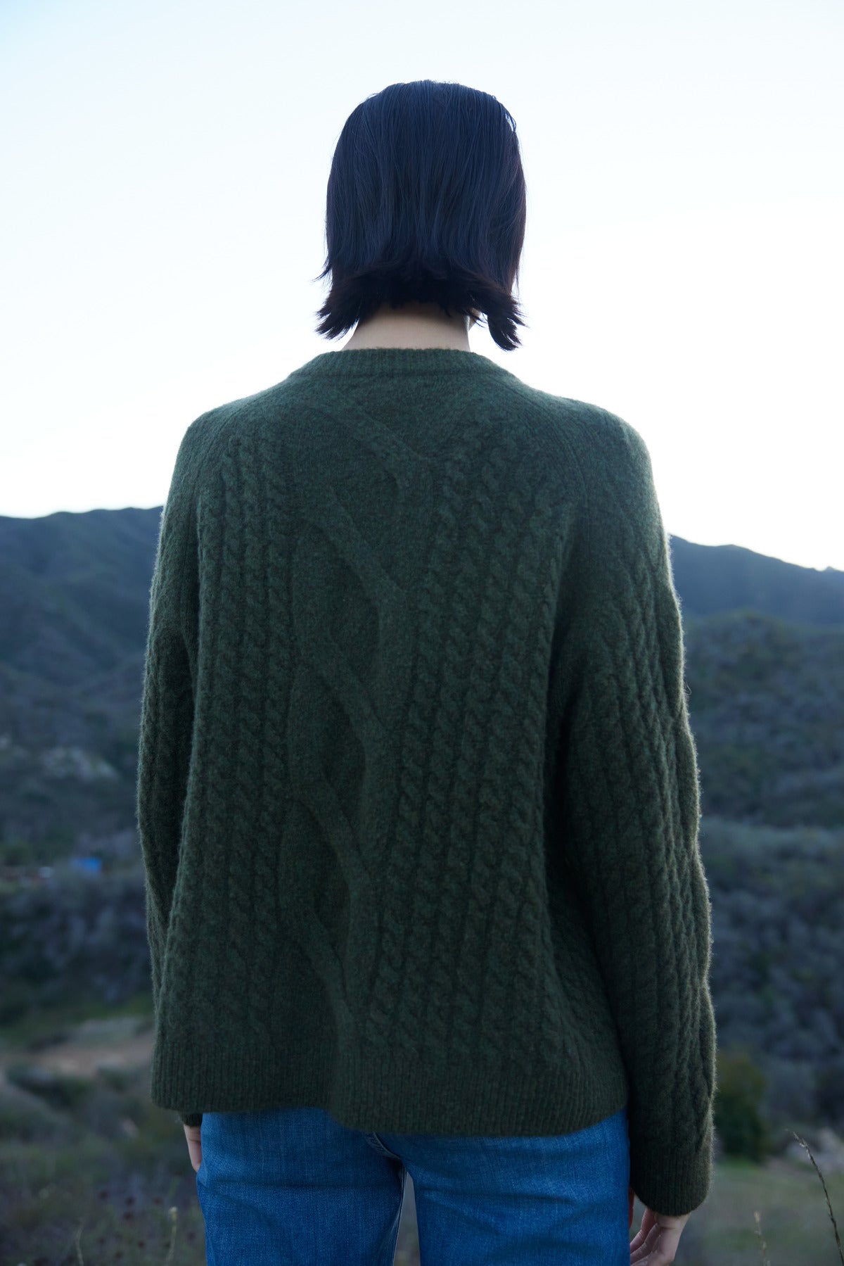 A person with short dark hair, wearing the BEACHWOOD CARDIGAN by Velvet by Jenny Graham, stands outdoors against a backdrop of mountains.-37665540440257