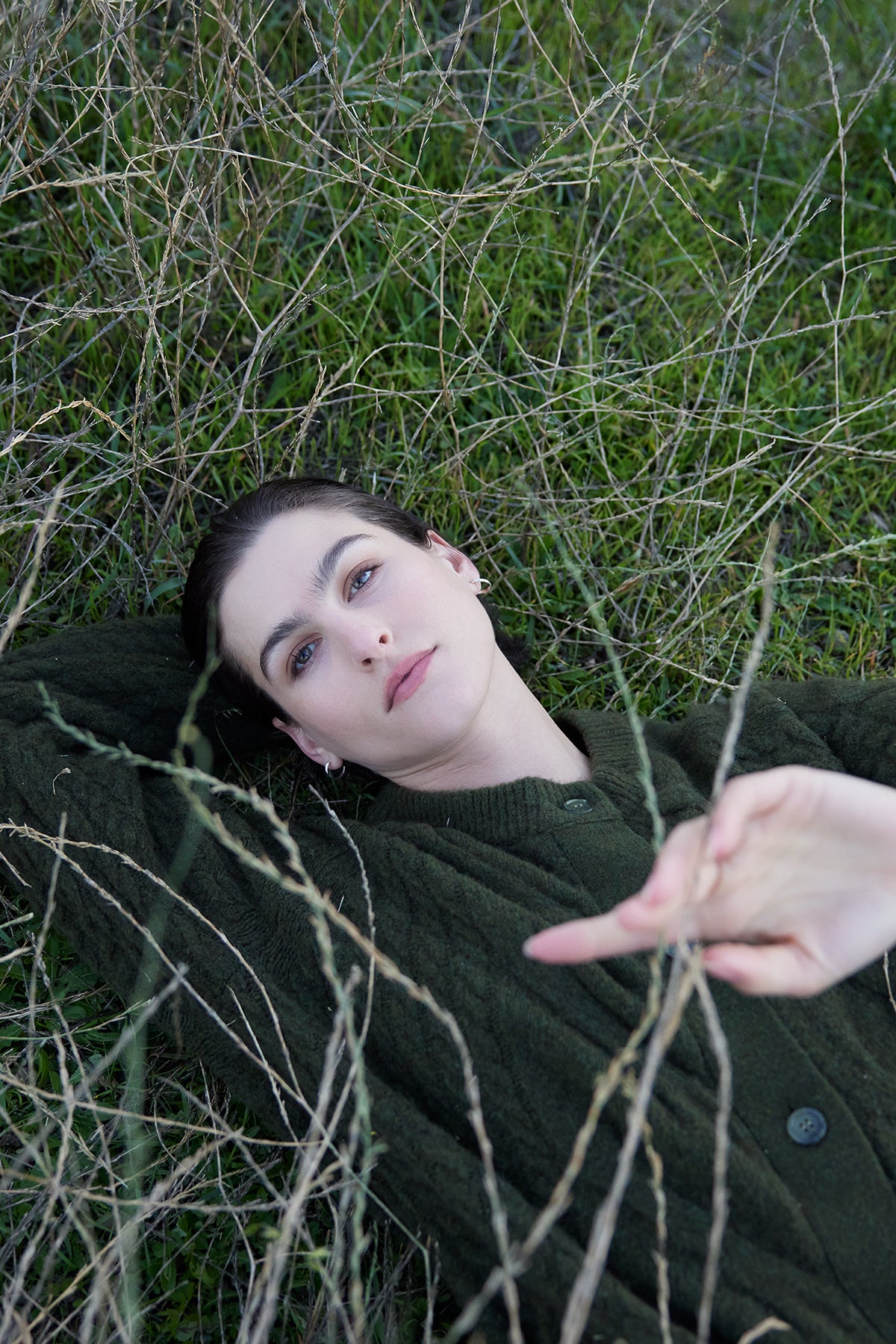   Person lying on grass, gazing upward, with arms partially visible through sparse dry branches, wearing the BEACHWOOD CARDIGAN by Velvet by Jenny Graham. 