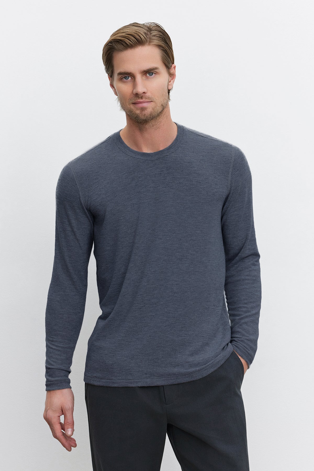   Dressed in the BECKER CREW by Velvet by Graham & Spencer, a man strikes a stylish pose against a plain white background, effortlessly showcasing everyday fashion in his cozy jersey gray long-sleeve shirt and dark pants. 