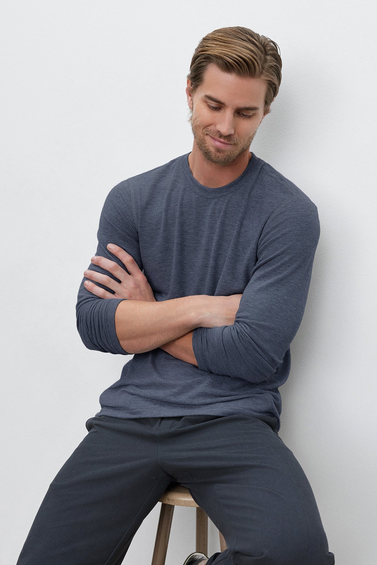   A man in a gray BECKER CREW by Velvet by Graham & Spencer and dark pants sits on a stool with arms crossed, looking down. 