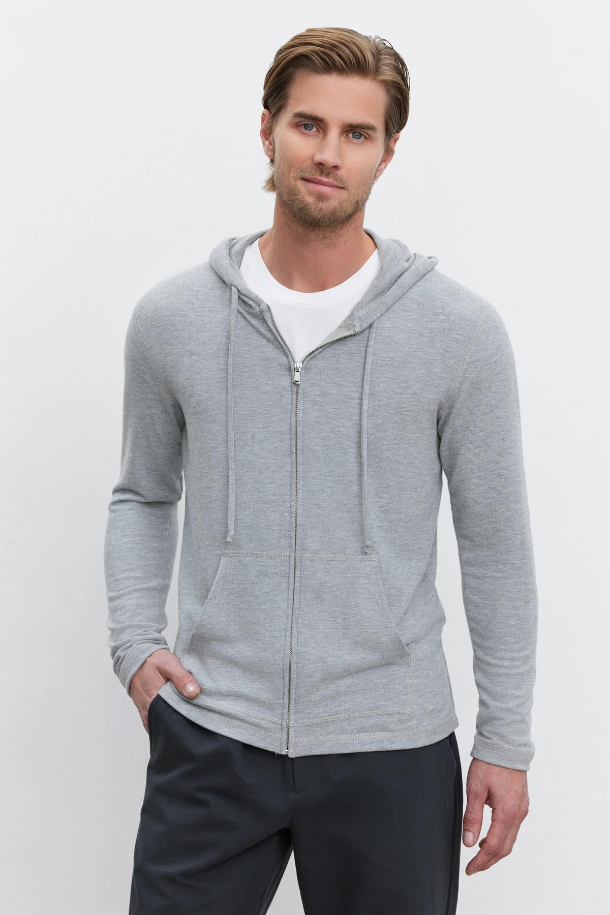   A man with short hair wearing a grey LOUDON HOODIE by Velvet by Graham & Spencer over a white t-shirt stands against a plain background, with one hand casually in his pocket. 