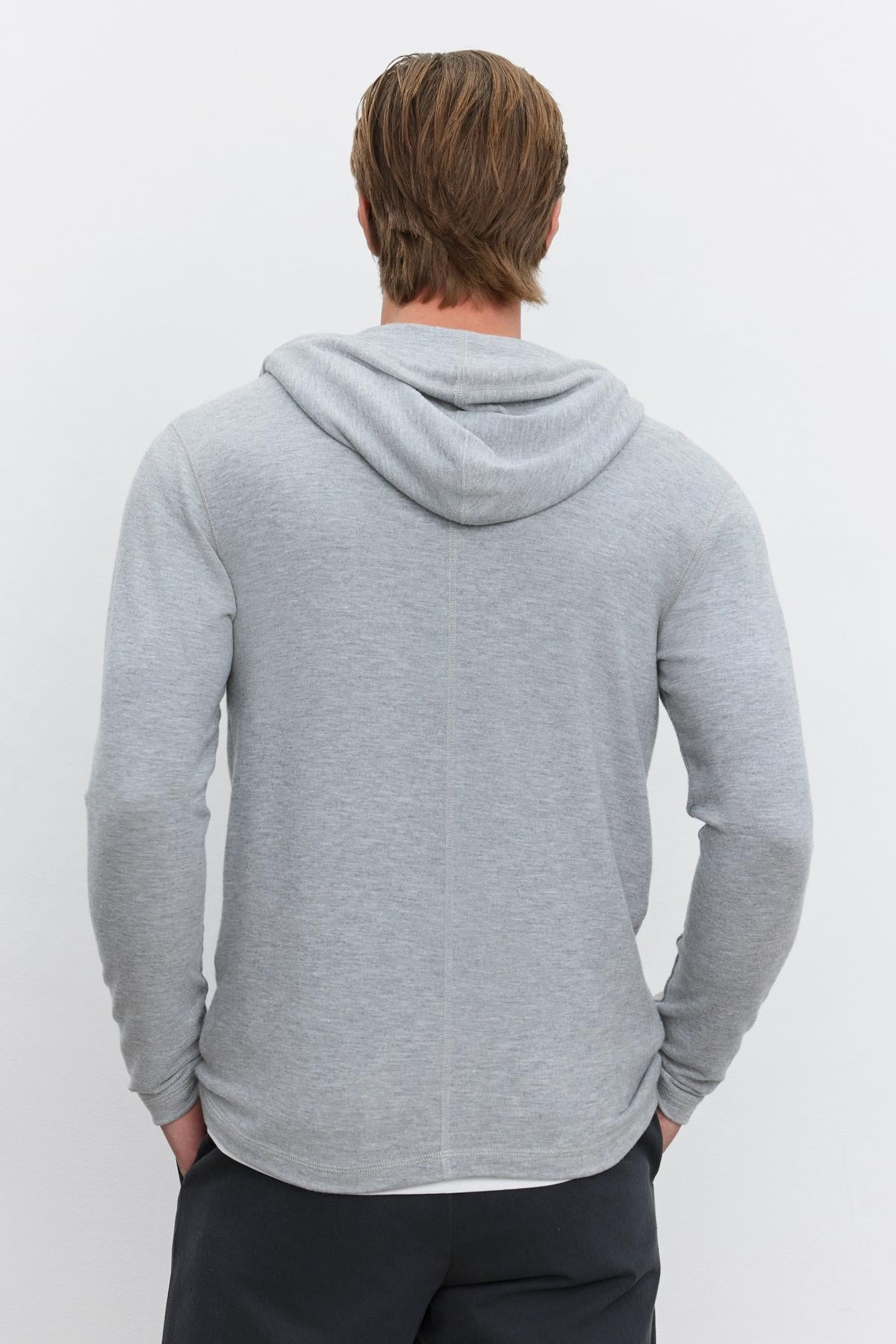   A person with light brown hair is standing with their back to the camera, wearing a cozy LOUDON HOODIE by Velvet by Graham & Spencer and dark pants. 