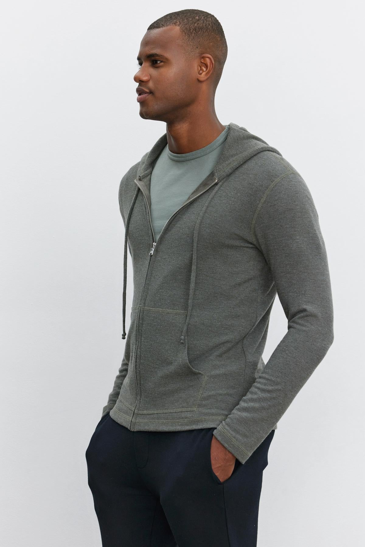   A man wearing a relaxed fit, gray LOUDON HOODIE by Velvet by Graham & Spencer and a green T-shirt stands with his hands in his pockets against a plain white background. 