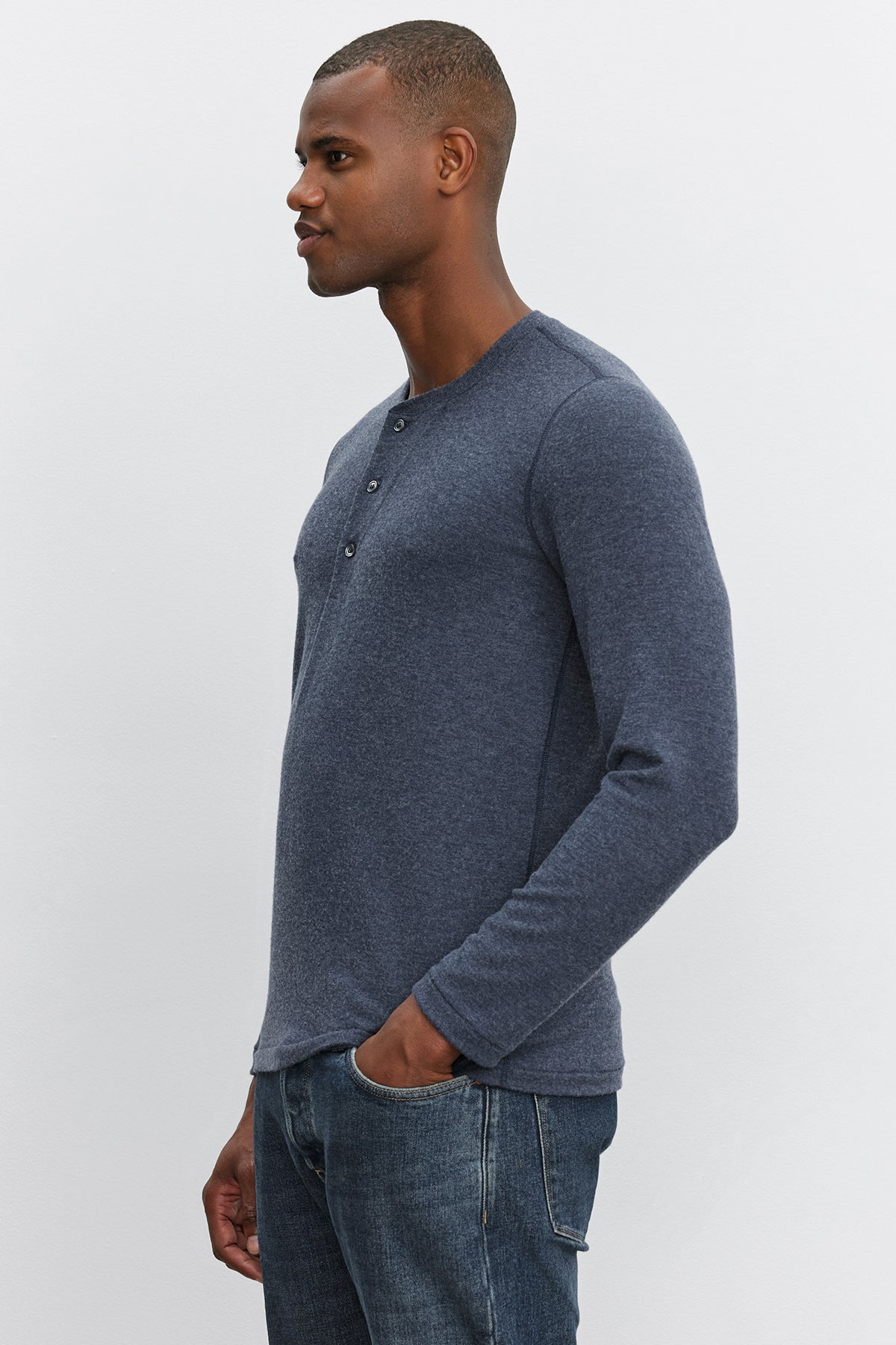   Side profile of a person standing with one hand in pocket, wearing a Velvet by Graham & Spencer RYLAND HENLEY shirt in cozy jersey blue and blue jeans against a plain white background. 