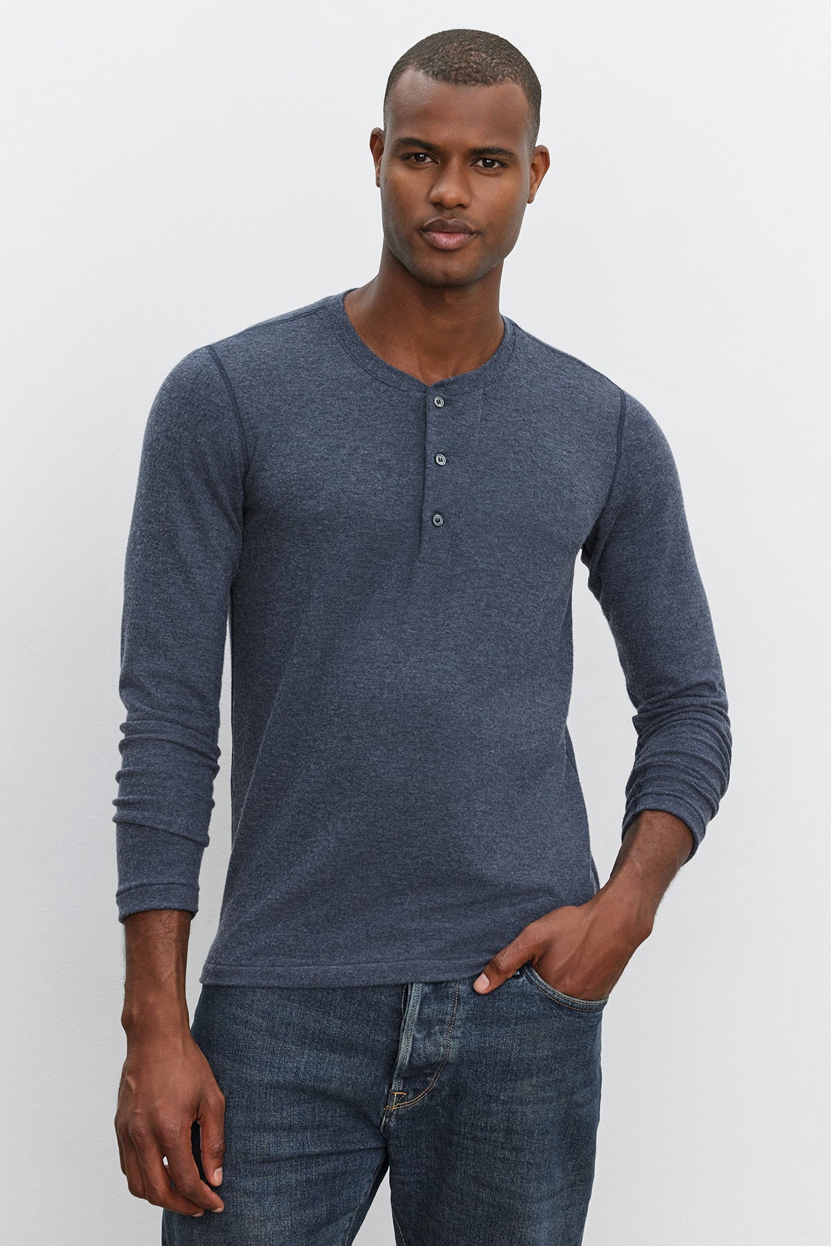 A person stands against a plain background wearing the RYLAND HENLEY, a blue 3-button shirt by Velvet by Graham & Spencer, paired with jeans and showcasing this versatile piece.-37893925667009