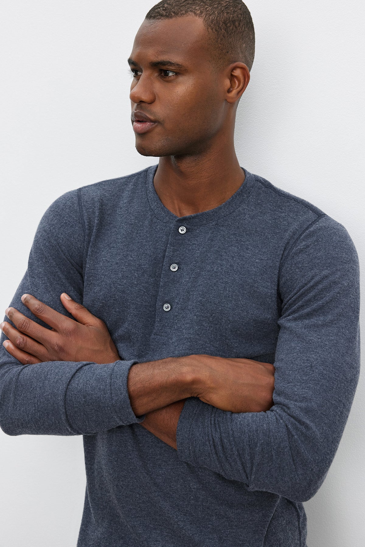   A man stands with arms crossed, looking to the side, wearing a RYLAND HENLEY long-sleeved, dark gray 3-button shirt from Velvet by Graham & Spencer against a plain white background. 