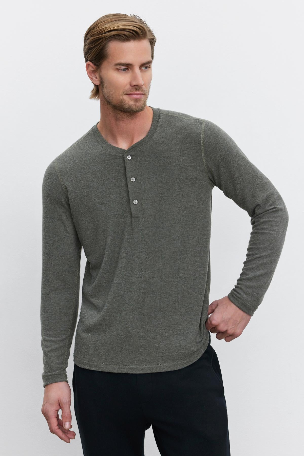   A man with short light brown hair is wearing the cozy RYLAND HENLEY in gray from Velvet by Graham & Spencer and black trousers. He gazes slightly to the side with his right hand resting on his hip, showcasing the versatility of this wardrobe piece. 