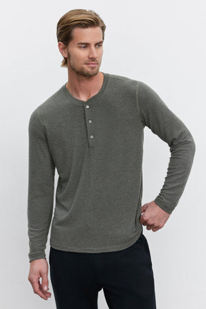 A man with short light brown hair is wearing the cozy RYLAND HENLEY in gray from Velvet by Graham & Spencer and black trousers. He gazes slightly to the side with his right hand resting on his hip, showcasing the versatility of this wardrobe piece.