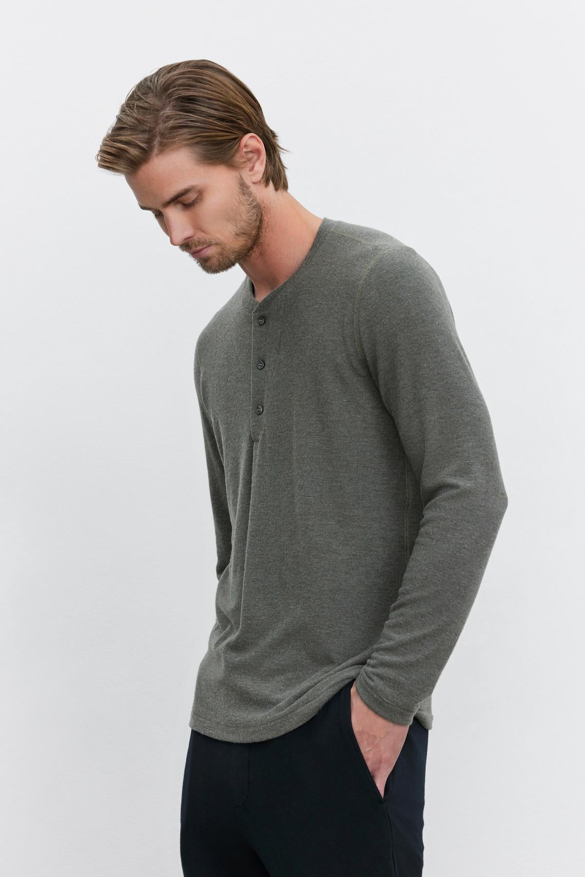   A man with short, brown hair is wearing a long-sleeved, grey RYLAND HENLEY shirt from Velvet by Graham & Spencer and black pants. He is looking downward with his left hand in his pocket against a plain white background. This versatile piece offers both comfort and style. 