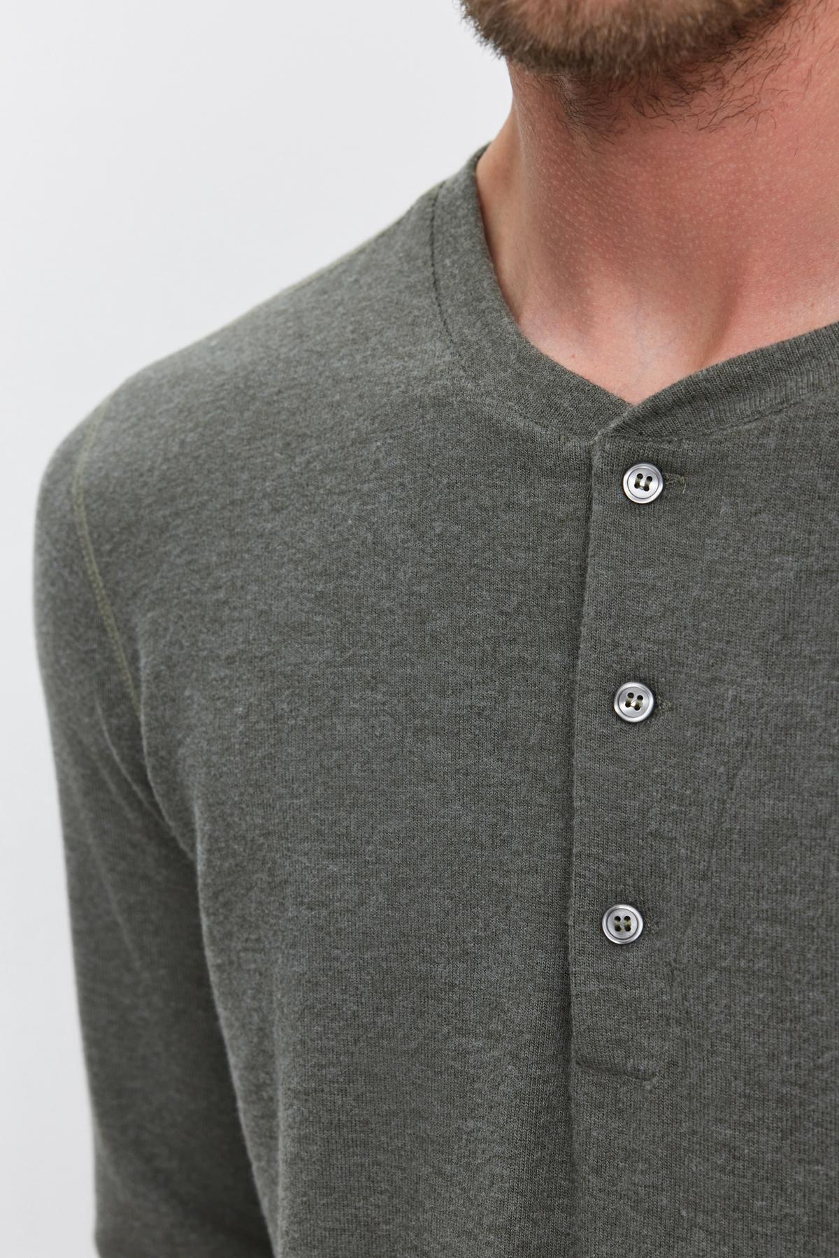   A close-up shows a person wearing the RYLAND HENLEY by Velvet by Graham & Spencer, a dark green, long-sleeved 3-button Henley shirt with three buttons fastened at the neckline. 