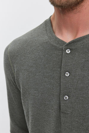 A close-up shows a person wearing the RYLAND HENLEY by Velvet by Graham & Spencer, a dark green, long-sleeved 3-button Henley shirt with three buttons fastened at the neckline.