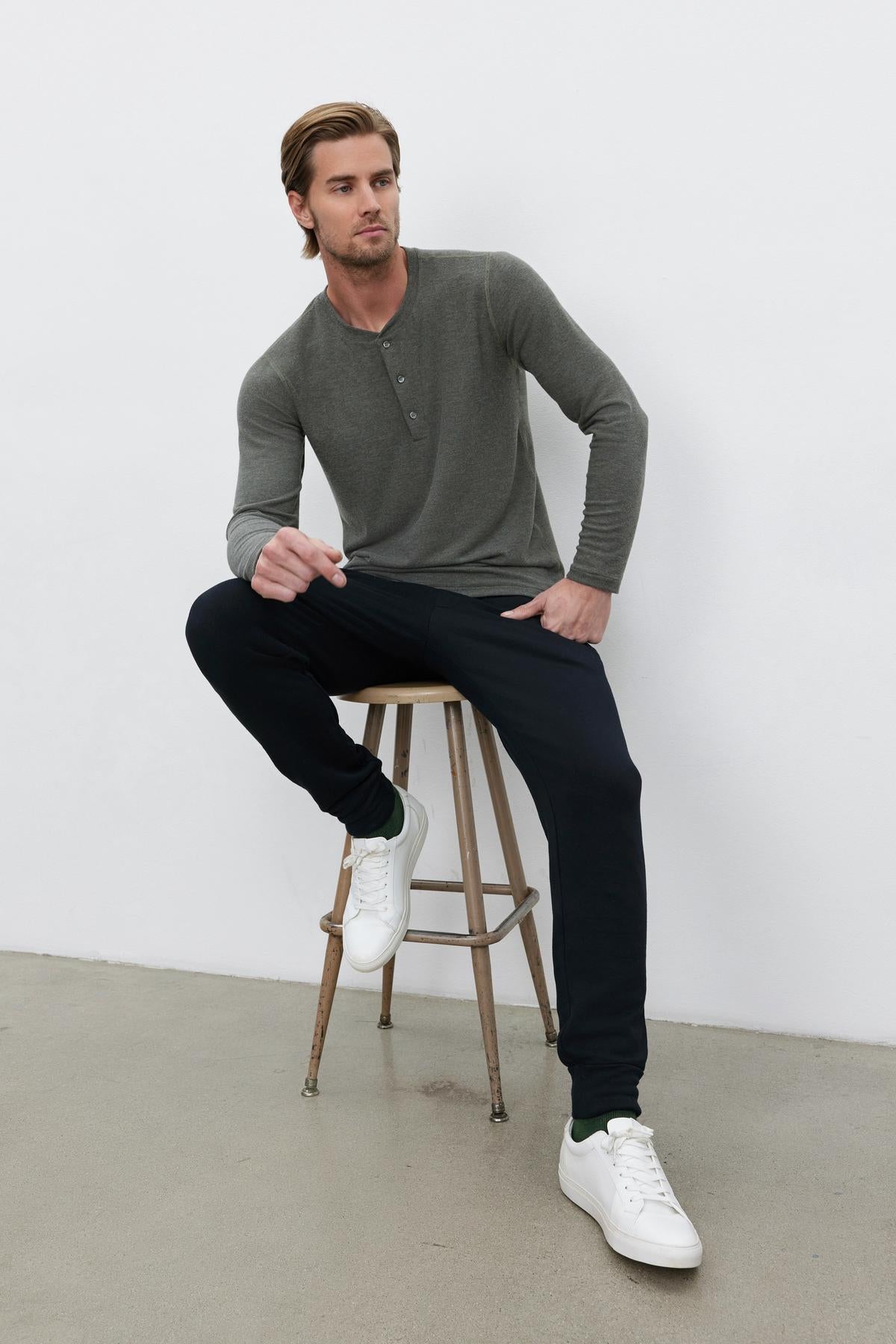 A man wearing a cozy RYLAND HENLEY in gray by Velvet by Graham & Spencer, black pants, and white sneakers sits on a wooden stool in a minimalistic indoor setting.-37759057723585