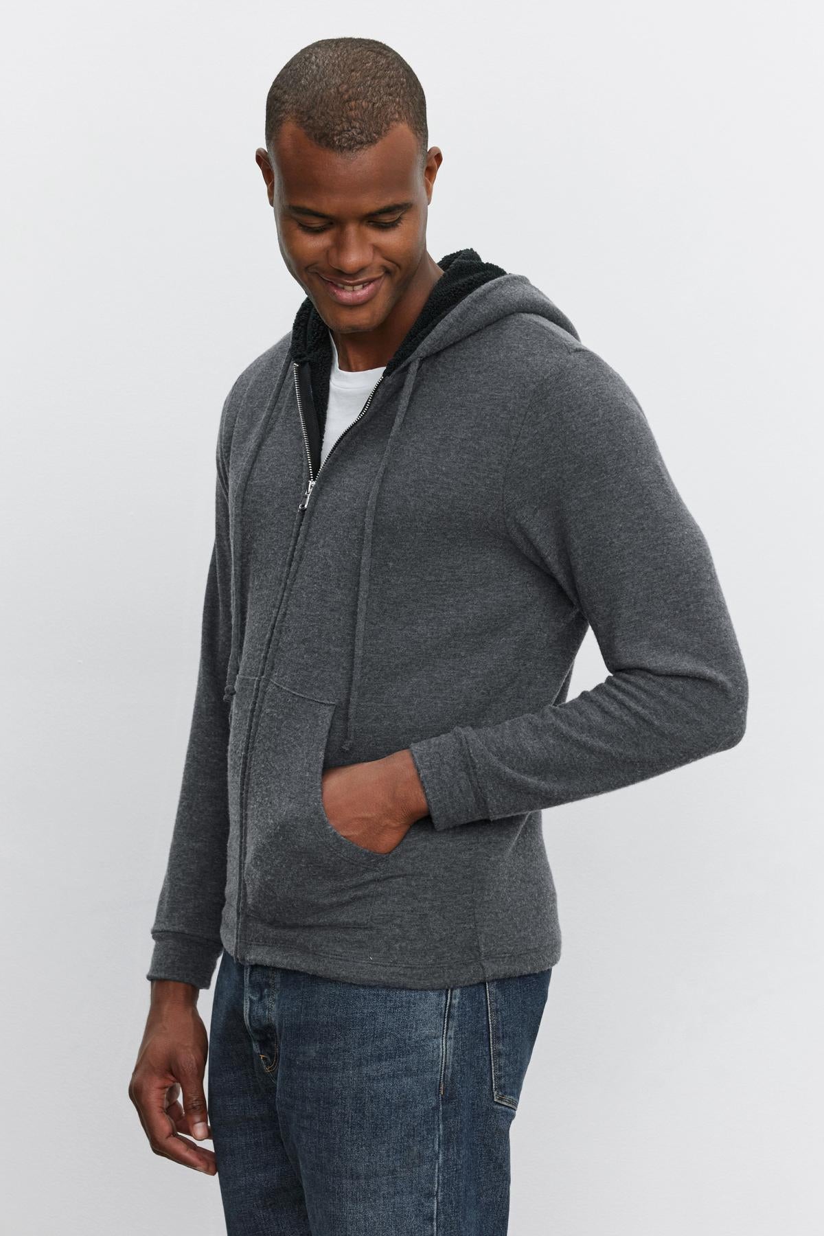   A person wearing a Velvet by Graham & Spencer SALVADORE HOODIE and jeans stands against a plain background, smiling and looking down. Their left hand is in their pocket. 