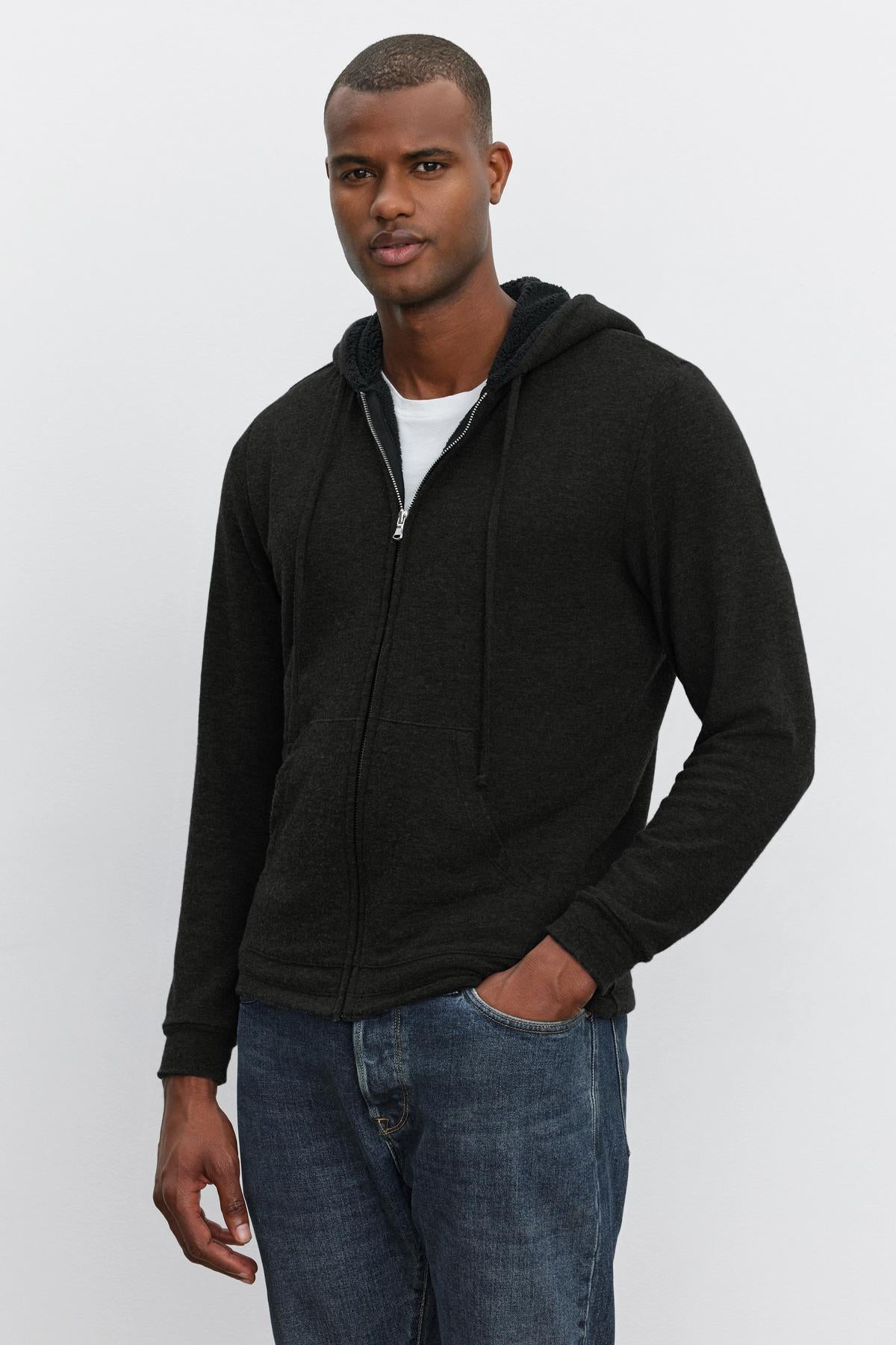 A man stands against a plain background wearing the SALVADORE HOODIE by Velvet by Graham & Spencer over a white t-shirt and blue jeans, with his left hand in his pocket.-37899375182017