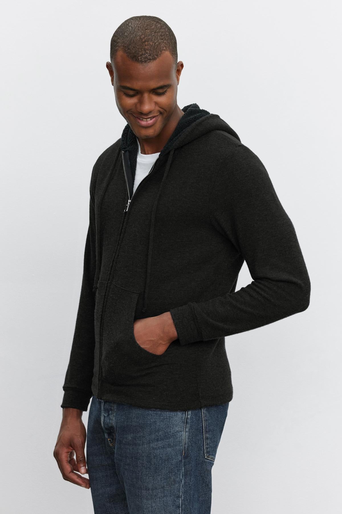 A man wearing a SALVADORE HOODIE by Velvet by Graham & Spencer, complete with faux sherpa lining, stands with one hand in his pocket over a white T-shirt, looking down and smiling.-37899375313089