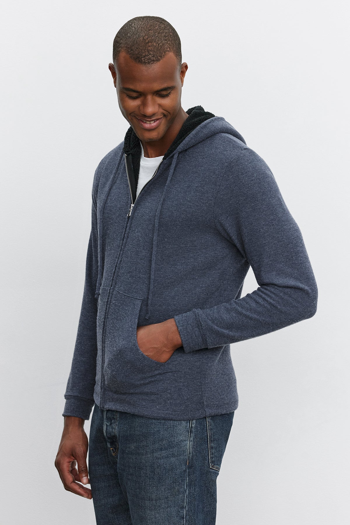   A person wearing the SALVADORE HOODIE by Velvet by Graham & Spencer and blue jeans stands with their hands in their pockets, looking down and smiling. 