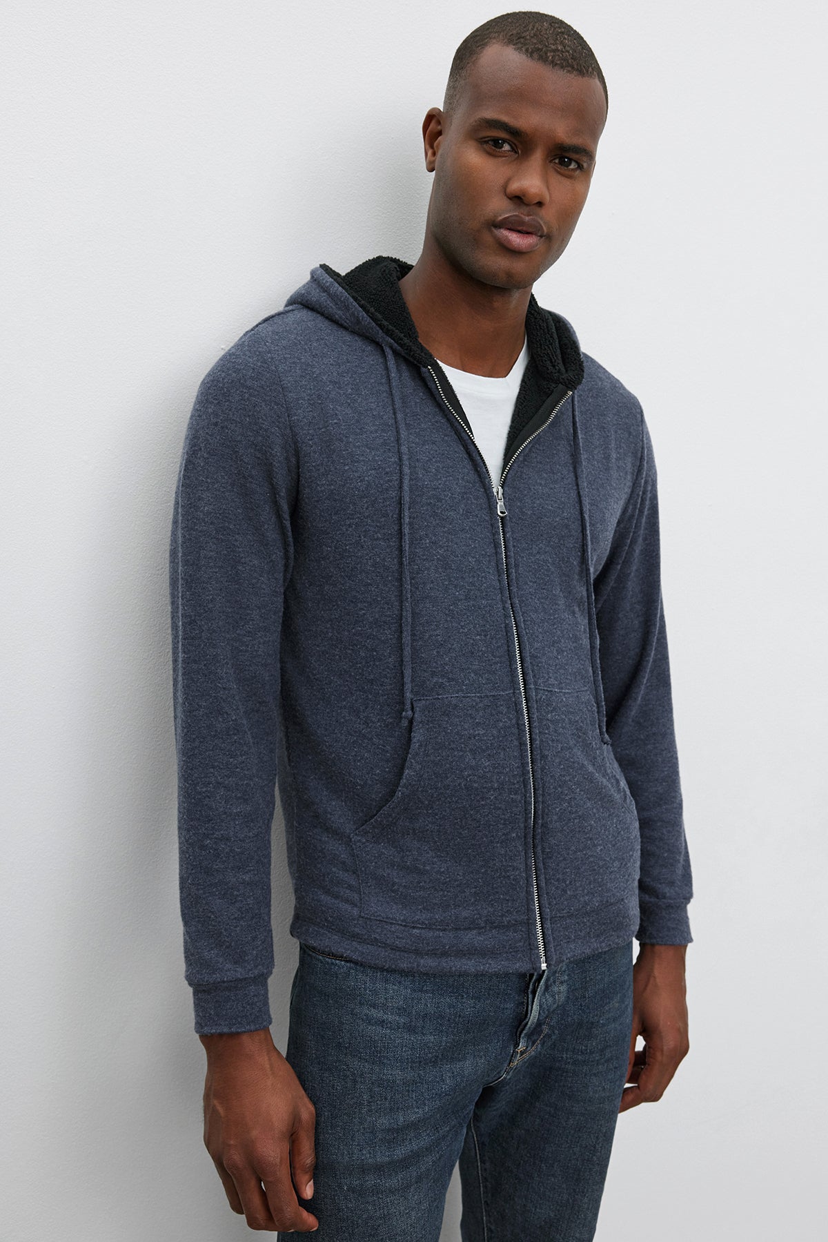   A man wearing a blue Velvet by Graham & Spencer SALVADORE HOODIE over a white t-shirt and blue jeans, standing against a plain wall. 