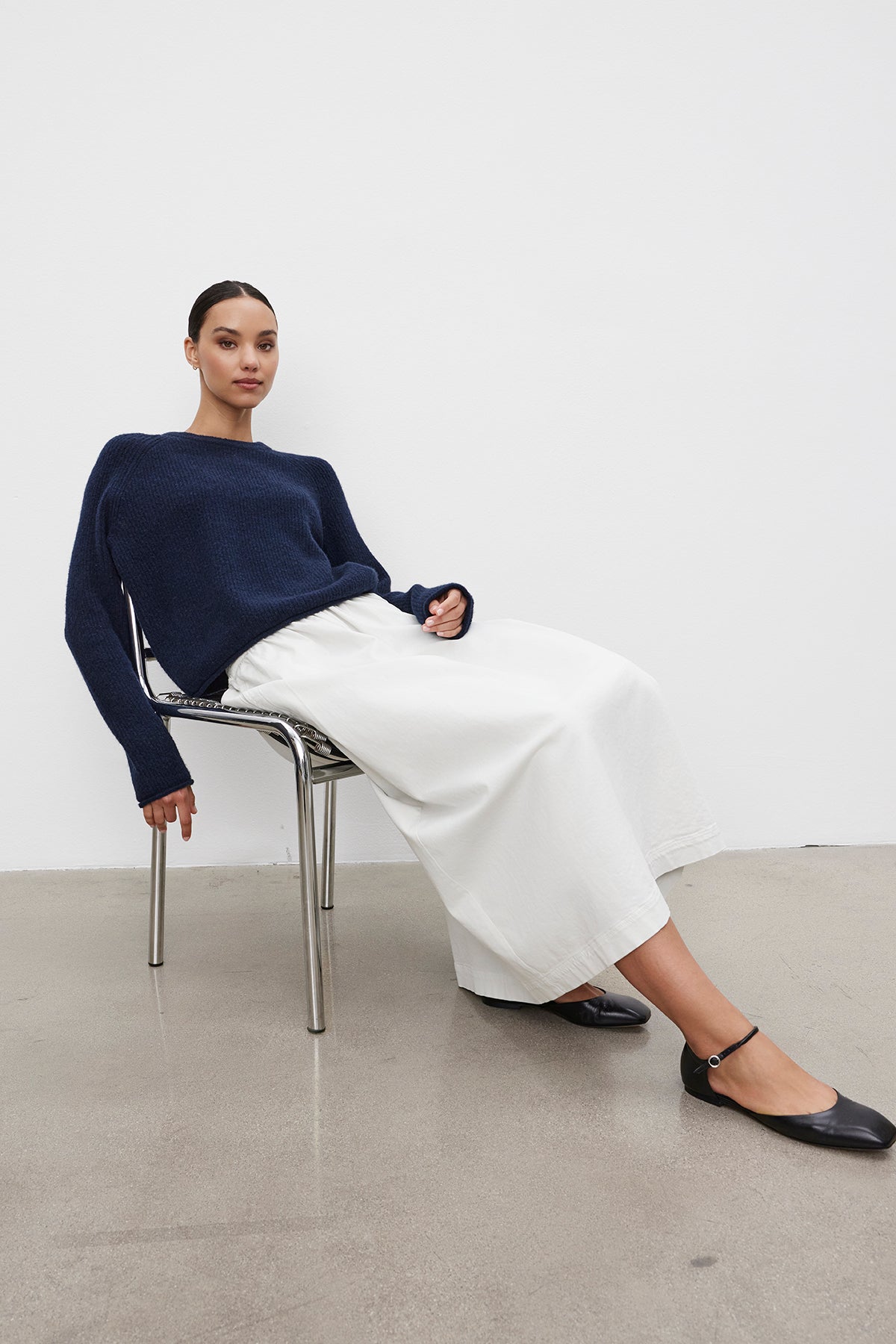 A person sitting on a chair against a white background, wearing a navy blue sweater, the SOPHIA SKIRT by Velvet by Graham & Spencer, and black flat shoes.-38116969185473