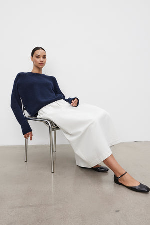 A person sitting on a chair against a white background, wearing a navy blue sweater, the SOPHIA SKIRT by Velvet by Graham & Spencer, and black flat shoes.