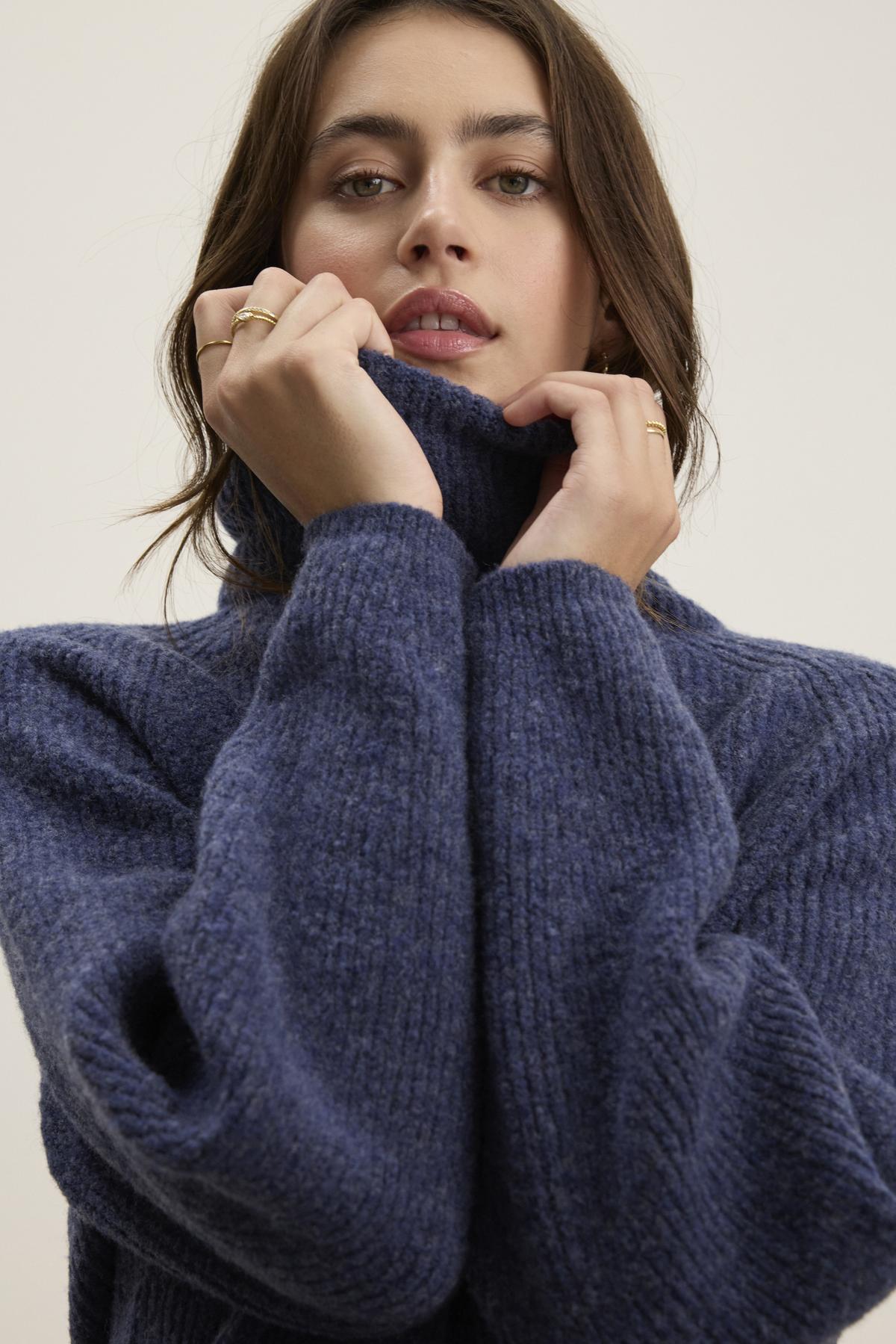   A person wearing the JUDITH TURTLENECK SWEATER by Velvet by Graham & Spencer pulls the cozy wool-blend blue collar close to their face, with their hands positioned near the chin. 