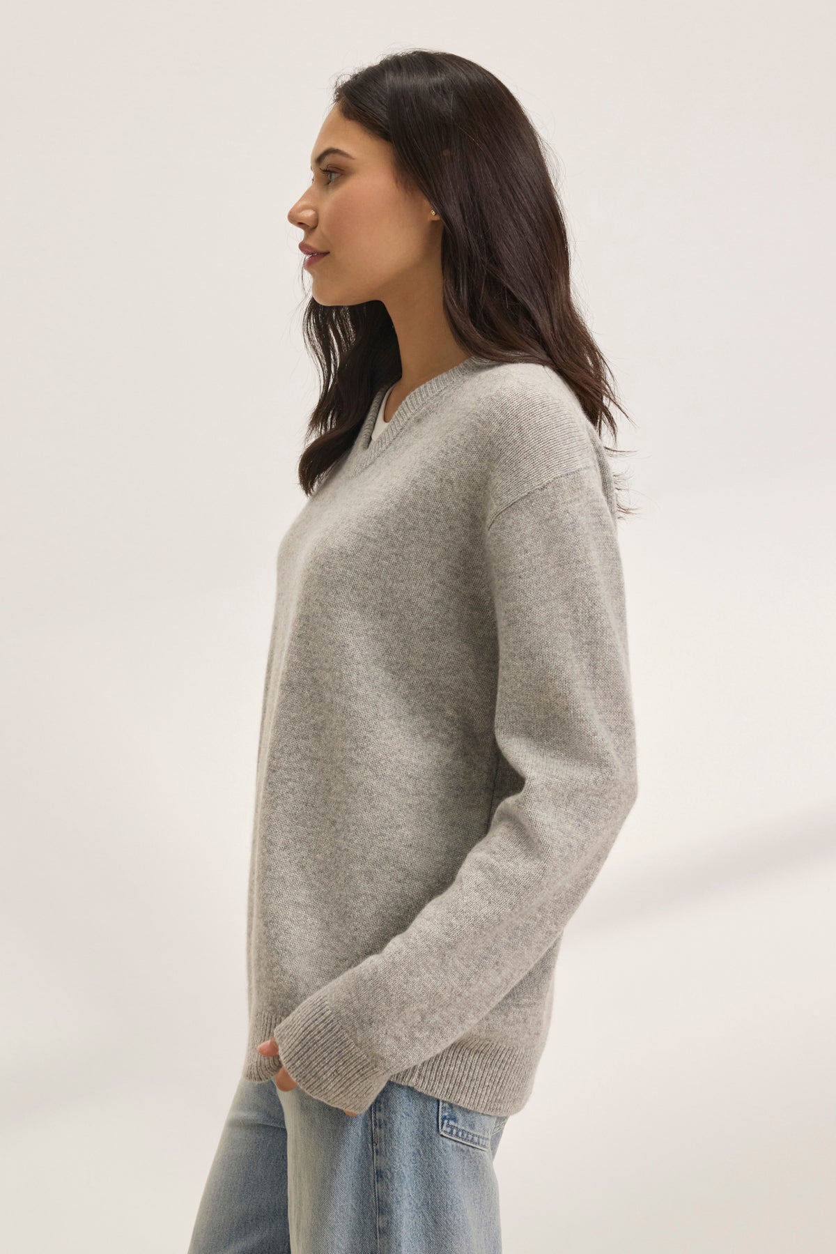   A person with long dark hair wears an oversized fit STEVIE WOOL CASHMERE SWEATER by Velvet by Graham & Spencer, paired with blue jeans, standing in profile against a plain background. 